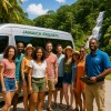 Group of nine happy tourists posing in front of a Jamaica Exquisite Transfer and Tours van with lush tropical trees and a waterfall in the background